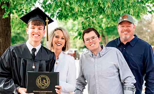 Picture of a family at graduation. The son on the far left next to his mom, brother and dad. Graduate is holding his diploma.
